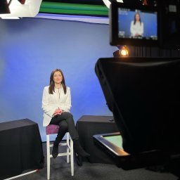 Marta Włodarczyk-Woźniczko Prawnik Przejęcia i finansowania spółek Corporate Governance Compliance Prawo bankowe - Kobieta w białej marynarce siedzi na krześle na tle niebieskiego ekranu w studio nagraniowym, widoczny monitor kamery z jej obrazem.