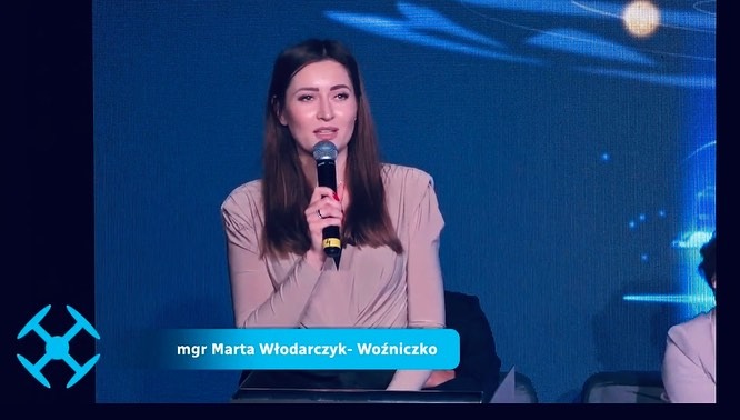 Kobieta, mgr Marta Włodarczyk-Woźniczko, przemawia do mikrofonu na tle niebieskiej ściany z logo. Prezentacja, konferencja, wydarzenie branżowe.