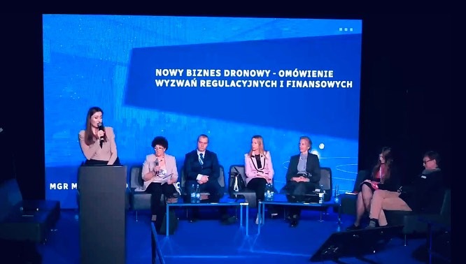 Konferencja o nowym biznesie dronowym: panel dyskusyjny z prelegentami na tle ekranu z tytułem. Kobieta przemawia zza mównicy, pozostali siedzą na krzesłach.