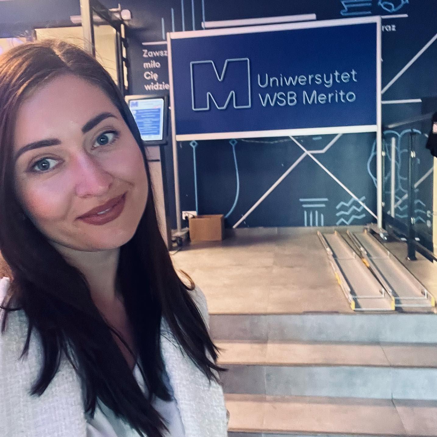Kobieta na tle logo Uniwersytetu WSB Merito, z rampą dla niepełnosprawnych w tle. Uśmiechnięta, w jasnej marynarce, patrzy w obiektyw.