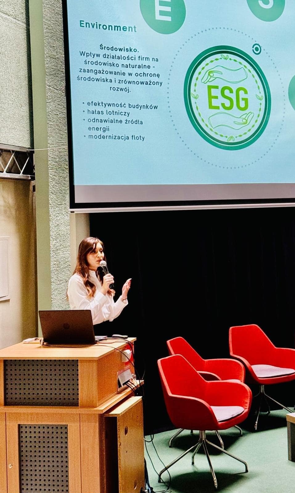 Prezentacja ESG: Kobieta z mikrofonem omawia wpływ firm na środowisko, efektywność budynków i odnawialne źródła energii. Na ekranie logo ESG i hasło 'Environment'.