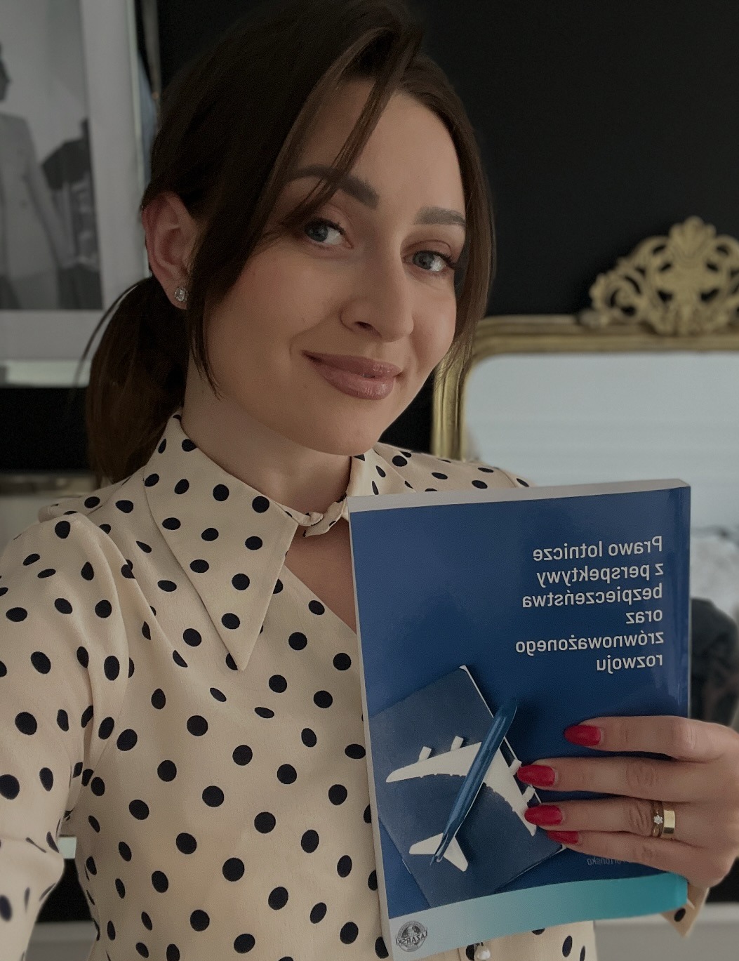 Uśmiechnięta kobieta w bluzce w grochy trzyma odwróconą książkę o prawie lotniczym z grafiką samolotu i paszportu. Czerwone paznokcie i pierścionki na palcach.