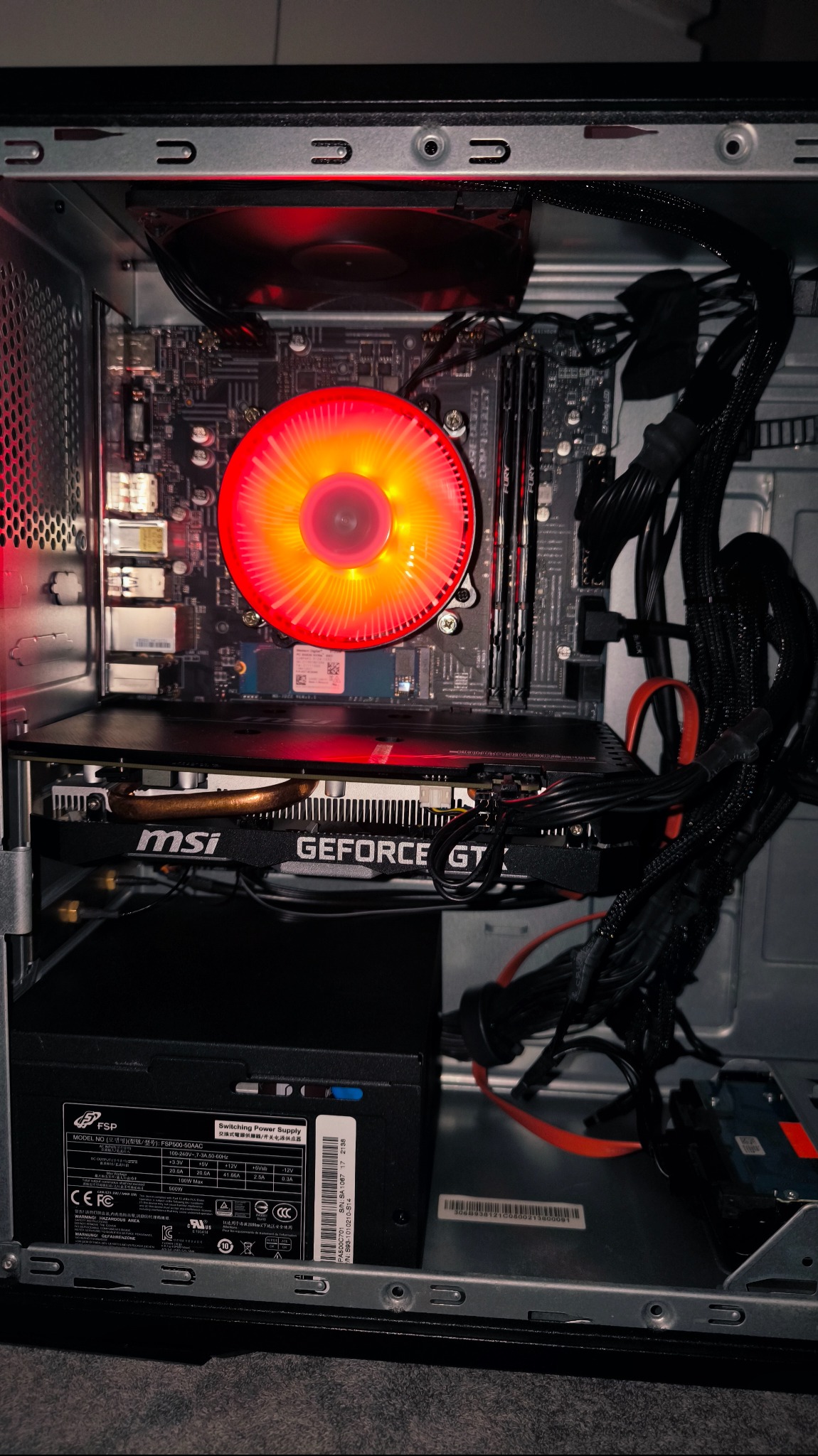 Wnętrze obudowy komputera stacjonarnego z podświetlanym na czerwono wentylatorem procesora, kartą graficzną MSI GeForce GT i zasilaczem FSP. Widoczne kable i płyta główna.