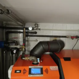 Pomarańczowy piec z czarnym kominem i rurami, na tle białych płytek. Widoczny manometr i oznaczenie 'Polski Produkt'.