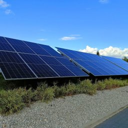 IQDOM24 Sp. z o.o. - Rząd paneli fotowoltaicznych z niebieskimi ogniwami, zamontowanych na trawiastym terenie z kamieniami, pod błękitnym niebem z chmurami. Widok z poziomu gruntu.