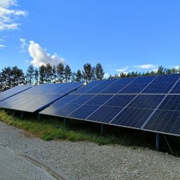 IQDOM24 Sp. z o.o. - Rząd paneli fotowoltaicznych na tle błękitnego nieba z chmurami i zieleni. Panele ustawione na gruncie, widoczne elementy konstrukcyjne. Ujęcie z dołu.