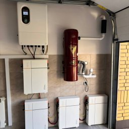 IQDOM24 Sp. z o.o. - Instalacja magazynu energii w garażu: biały inwerter Solax, trzy baterie Triple Power, brązowy odkurzacz centralny Cyclo Vac na ścianie z płytek. Widok na otwarte drzwi garażowe.