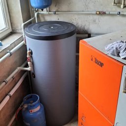 IQDOM24 Sp. z o.o. - Szary zasobnik ciepłej wody użytkowej obok pomarańczowego pieca Lazar Smart Fire, z niebieską butlą gazową w tle, w pomieszczeniu z instalacją hydrauliczną.