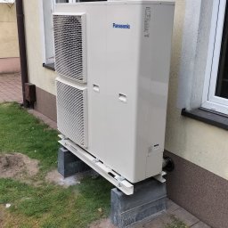 IQDOM24 Sp. z o.o. - Zewnętrzna jednostka pompy ciepła Panasonic, zamontowana na betonowych podstawach przy ścianie budynku z oknami. Widoczne elementy instalacji i trawnik.