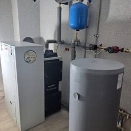 IQDOM24 Sp. z o.o. - Zestaw urządzeń grzewczych: piec, zbiornik buforowy i naczynie przeponowe w kotłowni. Instalacja wykonana w Gdańsku. Szare ściany i podłoga.