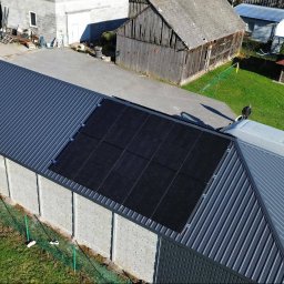 IQDOM24 Sp. z o.o. - Panel słoneczny na dachu budynku gospodarczego, widok z góry. Na drugim planie zabudowania gospodarcze i osoba stojąca obok białego busa.