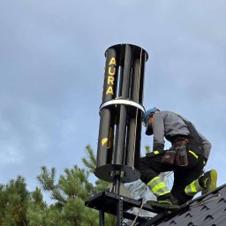 IQDOM24 Sp. z o.o. - Instalacja pionowej turbiny wiatrowej AURA na dachu przez montera w kombinezonie roboczym. Widok z dołu, pochmurne niebo w tle.