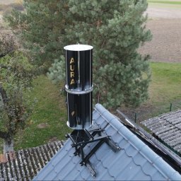 IQDOM24 Sp. z o.o. - Nietypowa, pionowa turbina wiatrowa AURA zamontowana na dachu budynku. Widok z góry, w tle drzewa i pole. Alternatywne źródło energii.