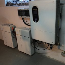 IQDOM24 Sp. z o.o. - Instalacja magazynu energii Solax Triple Power: dwa akumulatory i inwerter w pomieszczeniu z oknem. Widoczne okablowanie i elementy montażowe.