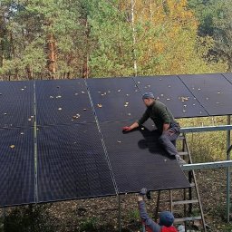 IQDOM24 Sp. z o.o. - Instalator czyści panele fotowoltaiczne z opadłych liści, stojąc na drabinie. W tle las. Widoczne mocowania paneli.