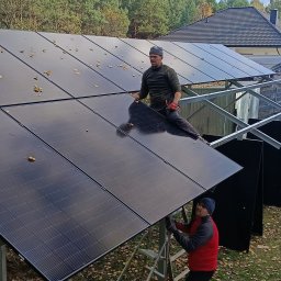 IQDOM24 Sp. z o.o. - Montaż paneli fotowoltaicznych na konstrukcji wsporczej przez dwóch instalatorów w jesiennej scenerii. Widoczne liście na panelach i dom w tle.