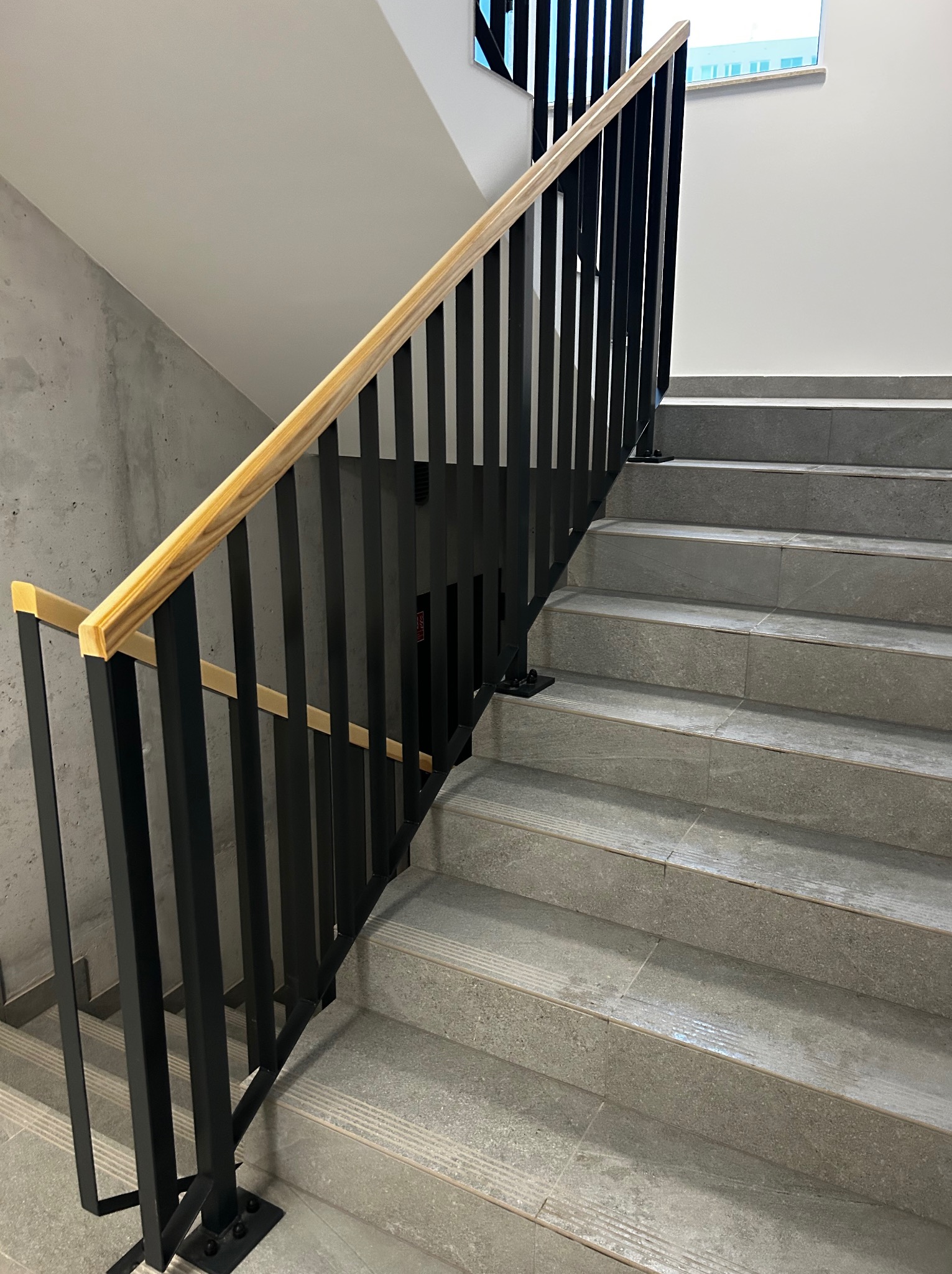 Nowoczesne schody z szarymi stopniami i czarną, metalową konstrukcją. Jasne, drewniane poręcze dodają ciepła surowemu wnętrzu. Minimalistyczny design.