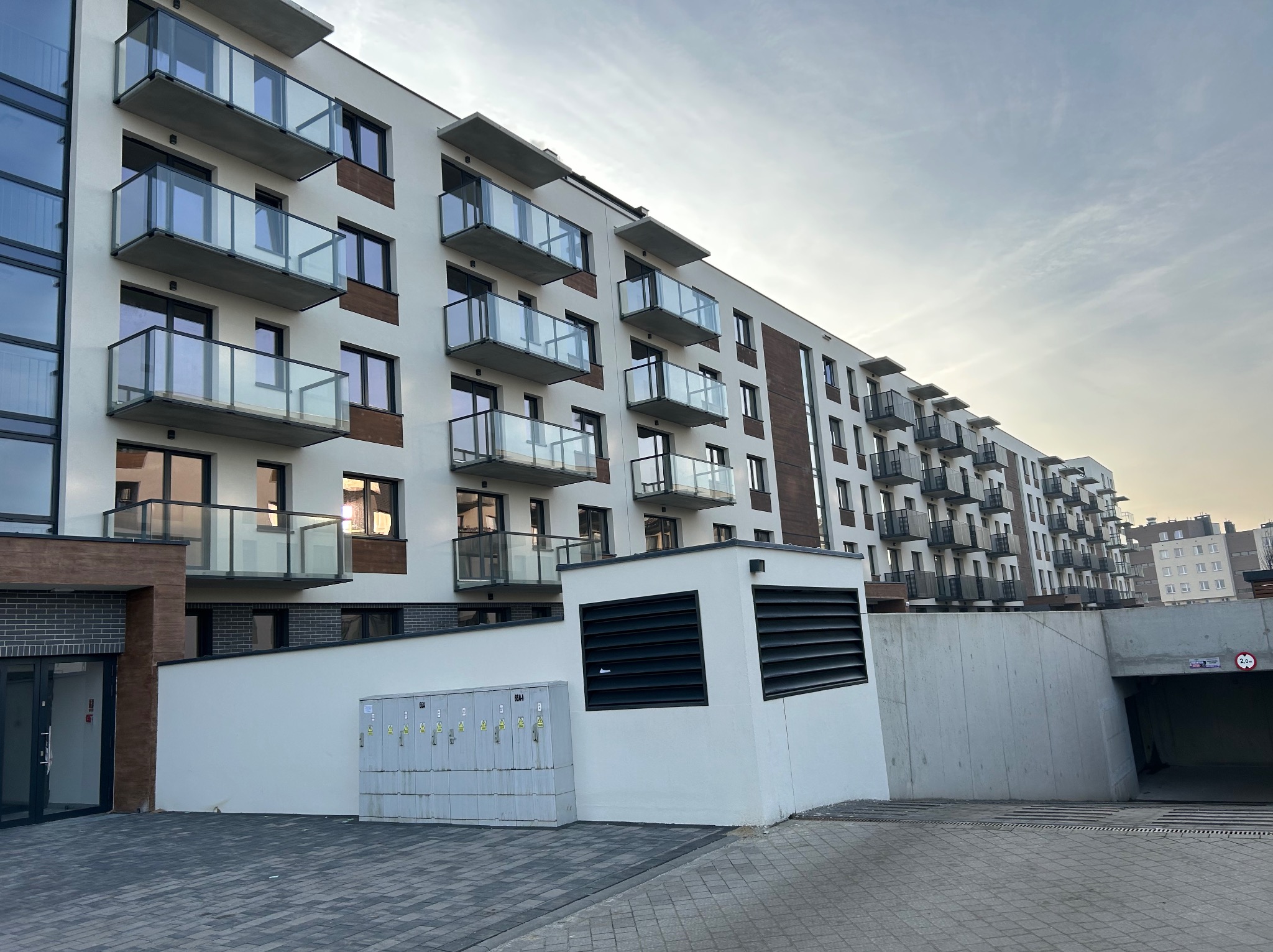 Nowoczesny budynek wielorodzinny z balkonami z przezroczystymi panelami. Widok z dołu na elewację z elementami drewna i szarej cegły. Wjazd do garażu podziemnego.