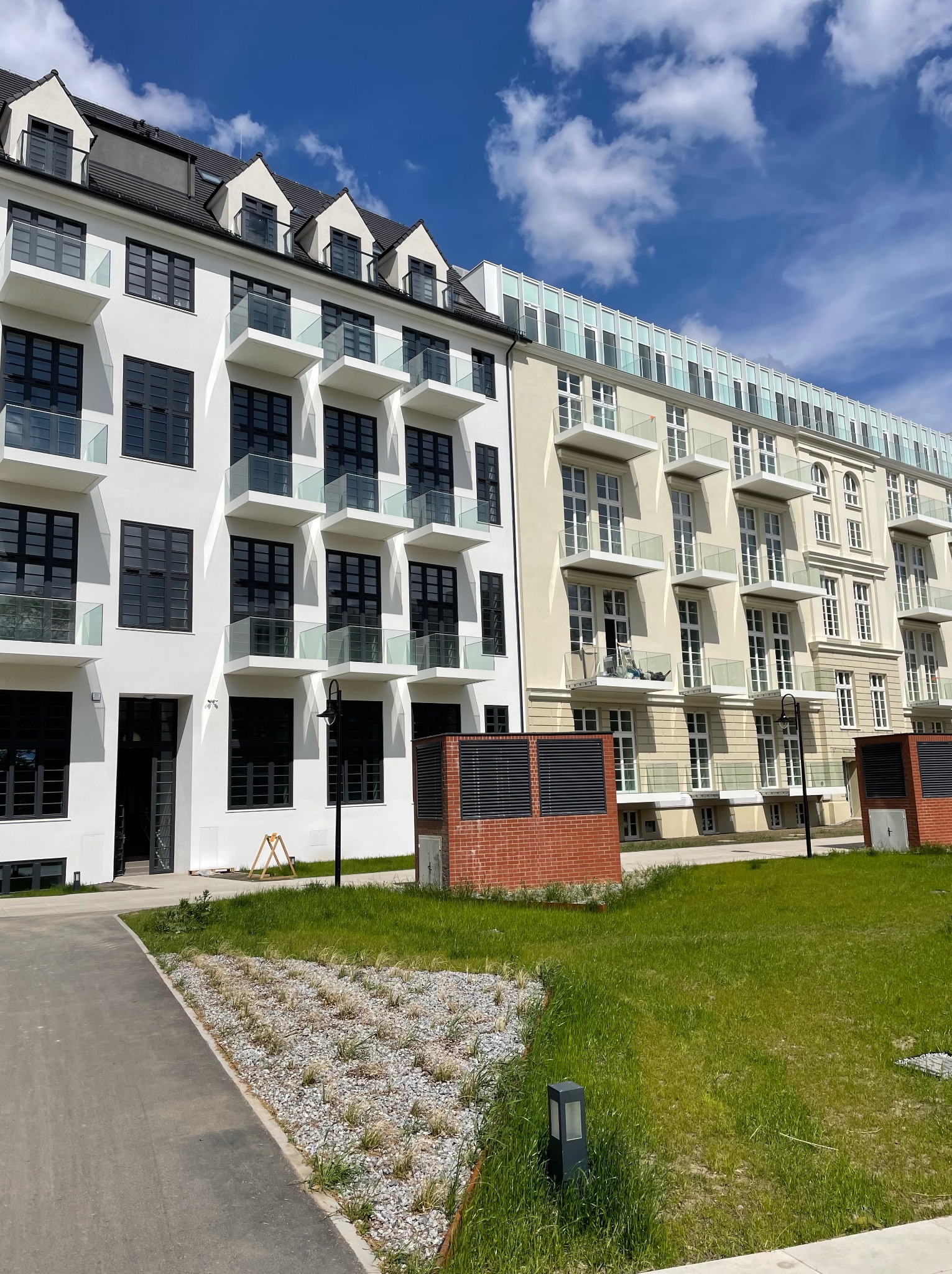 Nowoczesny budynek apartamentowy z białymi i beżowymi elewacjami, szklanymi balustradami balkonów i czarnymi ramami okiennymi, na tle błękitnego nieba.