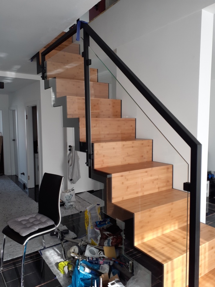 Nowoczesne schody z drewnianymi stopniami i szklaną balustradą z czarnymi elementami metalowymi. Widoczne niedokończone wykończenie wnętrza.