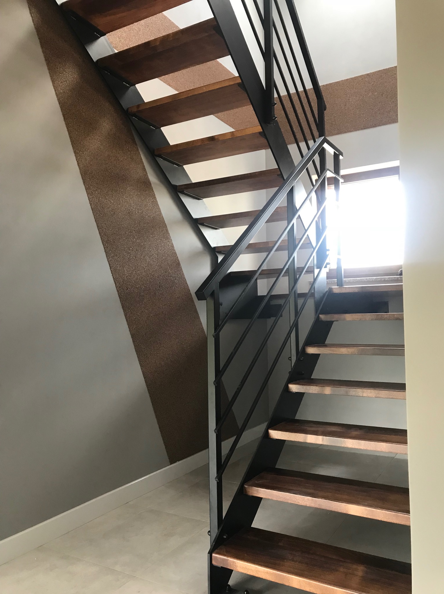 Nowoczesne schody metalowe z drewnianymi stopniami, czarne balustrady, jasne wnętrze z akcentem dekoracyjnym na ścianie. Ujęcie z dołu, widok na górę.