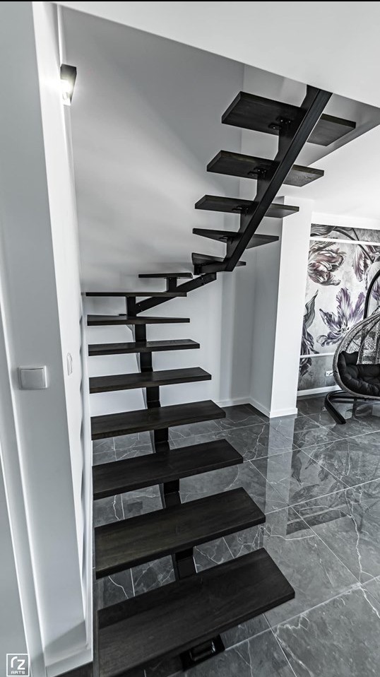 Nowoczesne schody metalowe z drewnianymi stopniami, minimalistyczny design, widok z dołu, szare płytki podłogowe, fotel wiszący w tle.