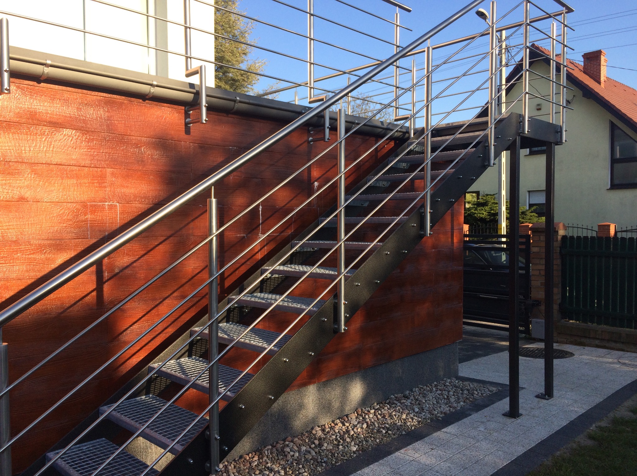 Zewnętrzne schody metalowe z ażurowymi stopniami i prostą, nowoczesną balustradą ze stali nierdzewnej, prowadzące do budynku o elewacji z desek w ciepłym odcieniu brązu.