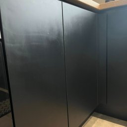 czystykąt - Matowe, czarne fronty szafek kuchennych z drewnianym blatem, widoczne smugi i zabrudzenia na powierzchni. Podłoga wyłożona jasnymi płytkami.