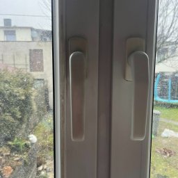 czystykąt - Brudne okno PCV z widocznymi zaciekami i osadami, uchwyt okienny w centrum kadru, zamazane tło z budynkiem i zielenią. Widok z bliska, słaba widoczność.