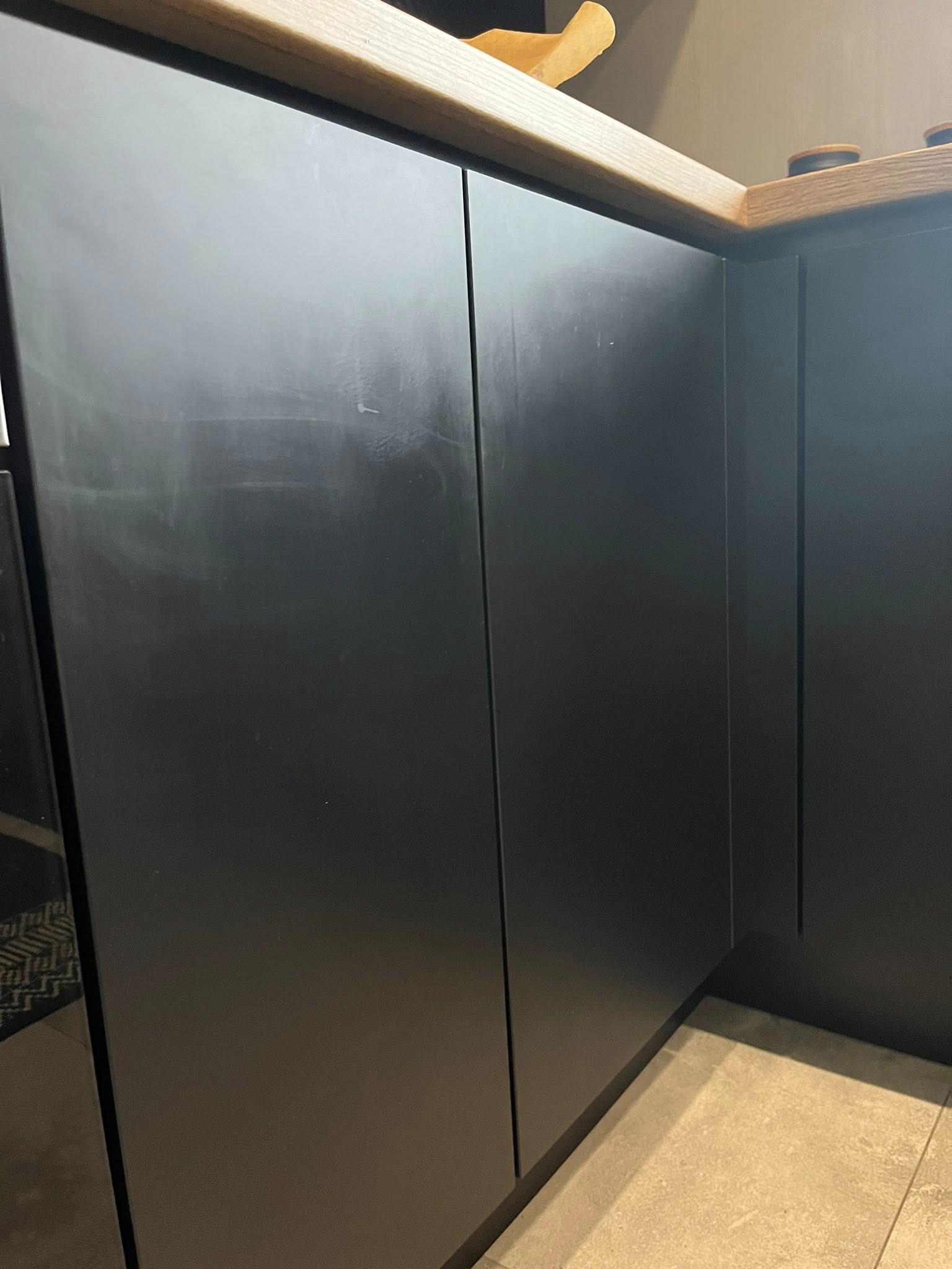 Matowe, czarne fronty szafek kuchennych z drewnianym blatem, widoczne smugi i zabrudzenia na powierzchni. Podłoga wyłożona jasnymi płytkami.