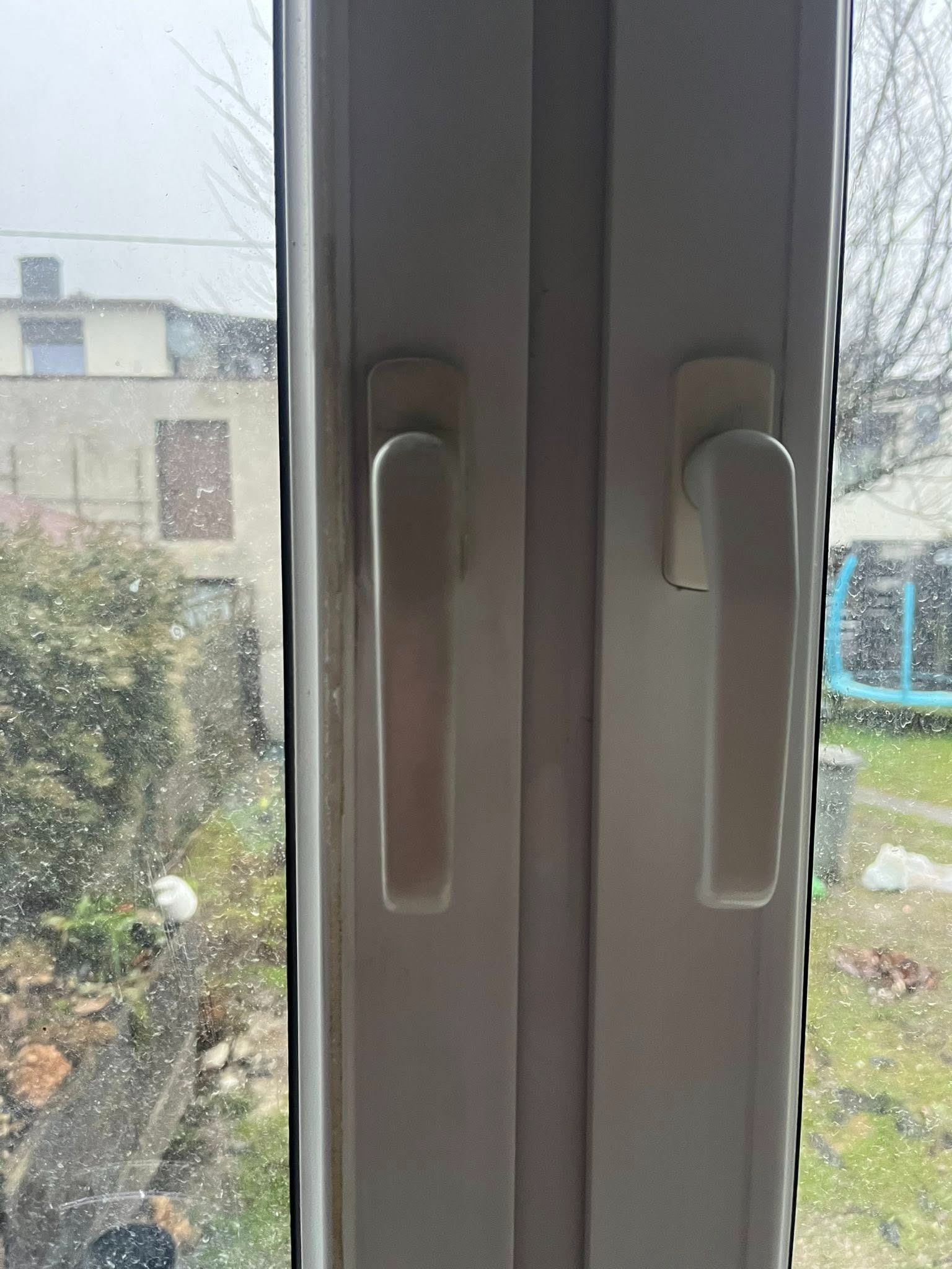 Brudne okno PCV z widocznymi zaciekami i osadami, uchwyt okienny w centrum kadru, zamazane tło z budynkiem i zielenią. Widok z bliska, słaba widoczność.