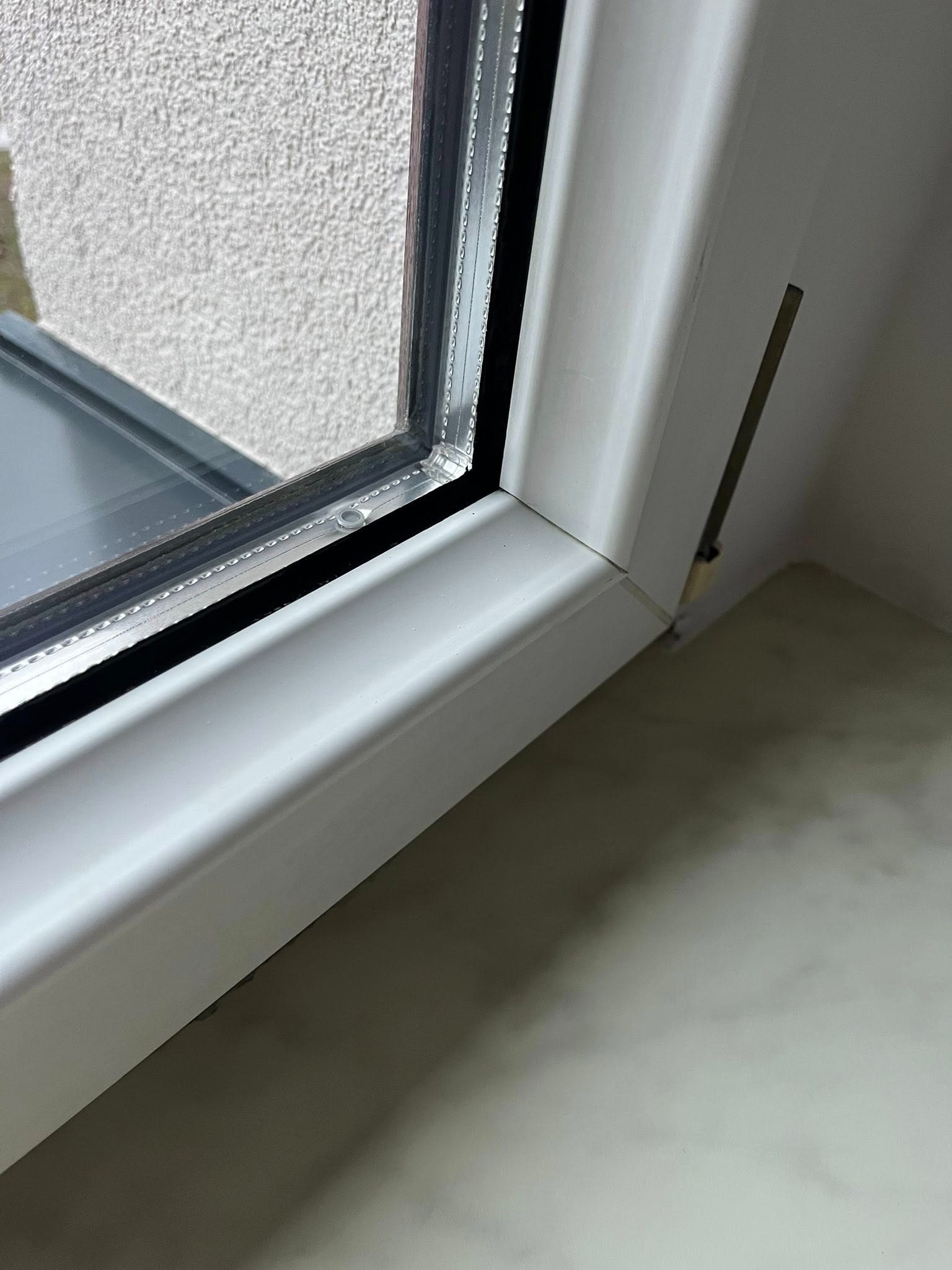 Szczegółowe ujęcie narożnika okna PCV z widocznymi uszczelkami i fragmentem ściany o chropowatej fakturze. Widoczne ślady po czyszczeniu.