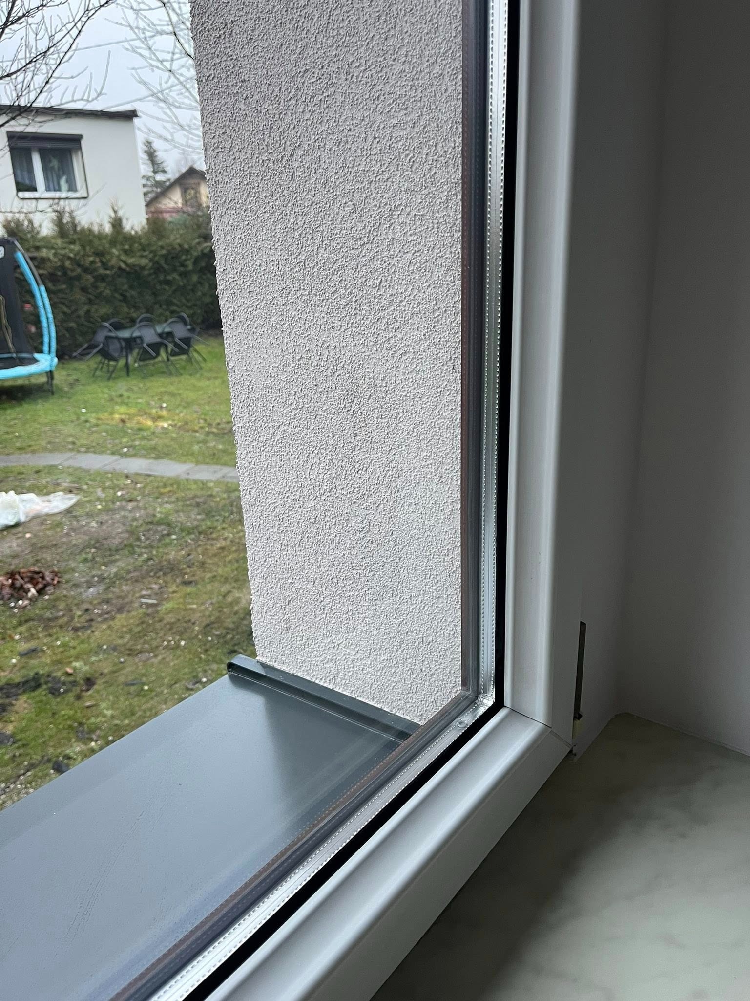 Nowe, białe okno PCV z widokiem na ogród z trampoliną i meblami ogrodowymi. Szary parapet wewnętrzny. Teksturowana, jasna elewacja budynku.