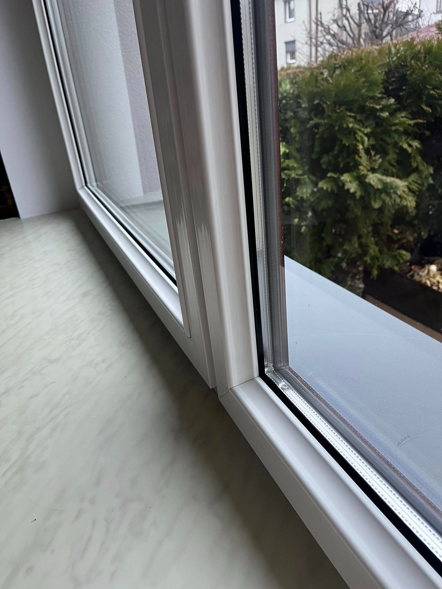 Białe, plastikowe okno z widokiem na zieleń i budynek. Widoczny parapet z marmurkowym wzorem. Ujęcie z bliska, skupione na detalu ramy okiennej i widoku za szybą.