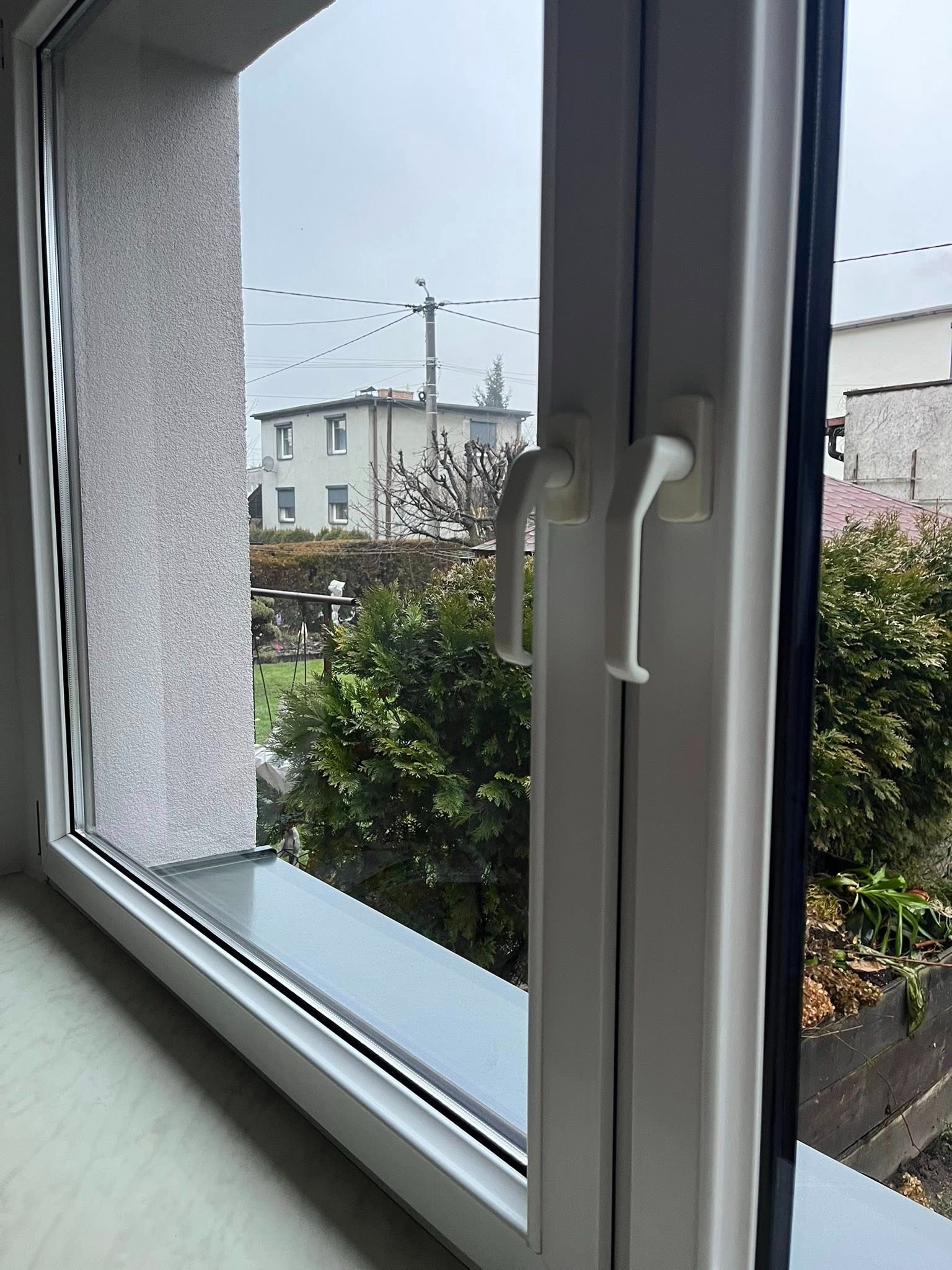 Otwarty, biały plastikowy okno z widokiem na ogród i dom w pochmurny dzień. Widoczne detale ramy okiennej i uchwytów.