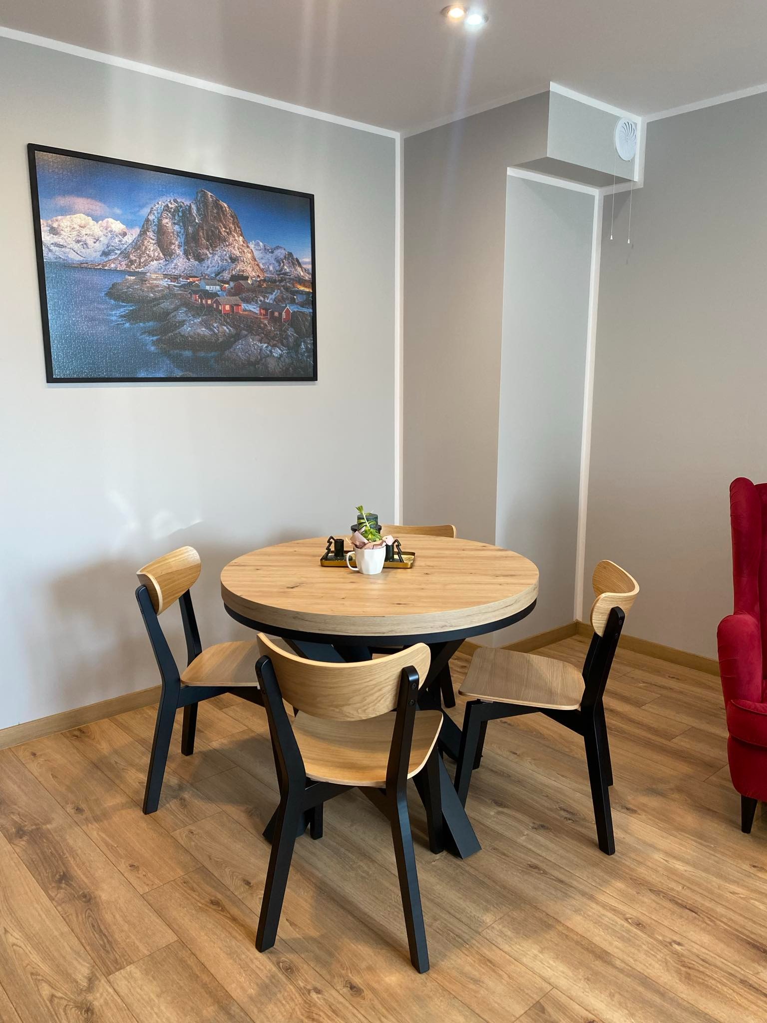 Jasne wnętrze z okrągłym stołem i krzesłami na drewnianej podłodze. Na ścianie wisi obraz z górskim krajobrazem. Widoczny fragment czerwonego fotela.