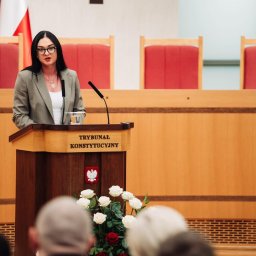 Adwokat Dr Marlena Paszko Kancelaria Adwokacka - Kobieta w okularach przemawia z mównicy Trybunału Konstytucyjnego, w tle flagi Polski i krzesła. U dołu widoczne kwiaty i fragmenty głów słuchaczy.