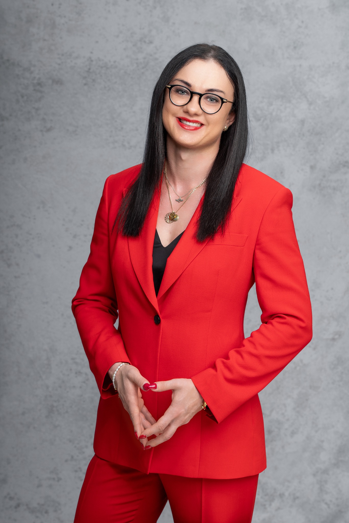 Portret uśmiechniętej adwokatki w czerwonym garniturze i okularach na tle szarej ściany. Złożone dłonie, widoczne detale biżuterii.