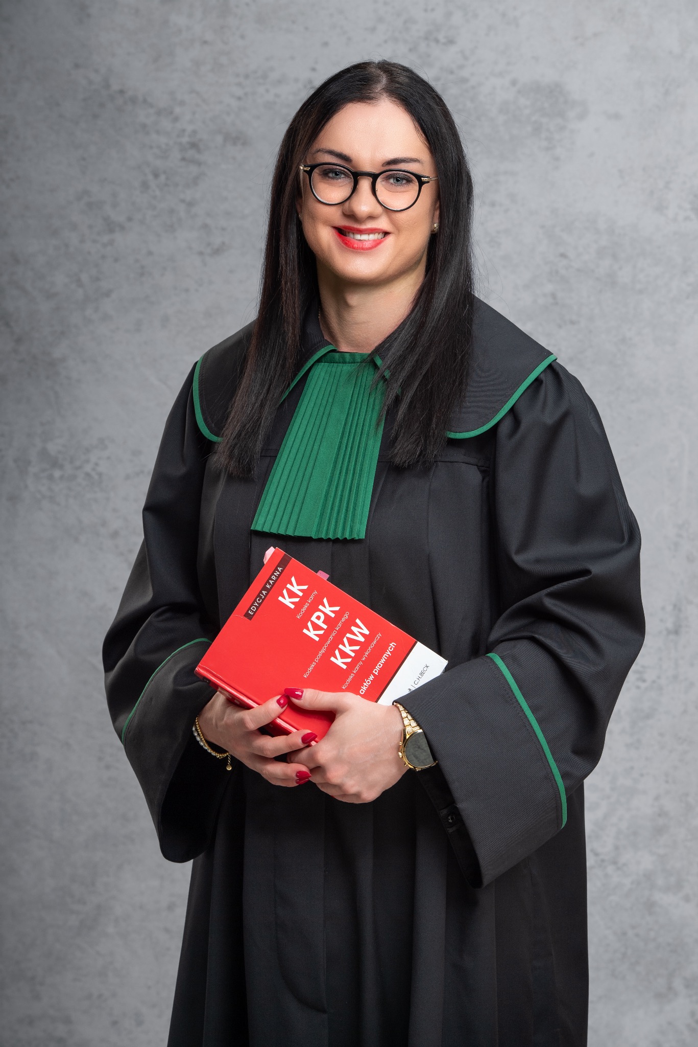 Uśmiechnięta adwokat w todze, trzymająca czerwoną książkę z napisem 'KK KPK KKW', na szarym tle. Widoczne okulary, zegarek i manicure.