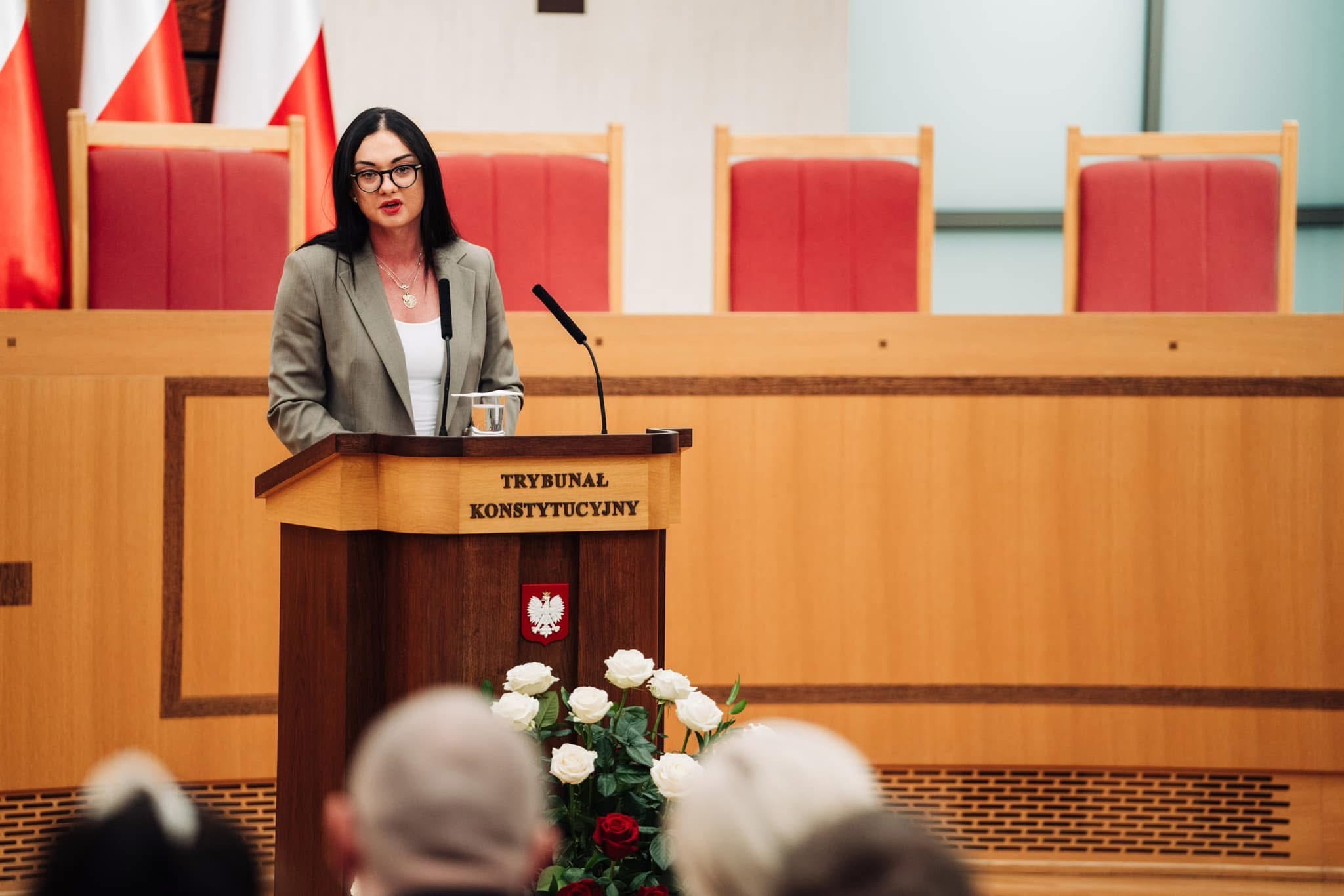Kobieta w okularach przemawia z mównicy Trybunału Konstytucyjnego, w tle flagi Polski i krzesła. U dołu widoczne kwiaty i fragmenty głów słuchaczy.