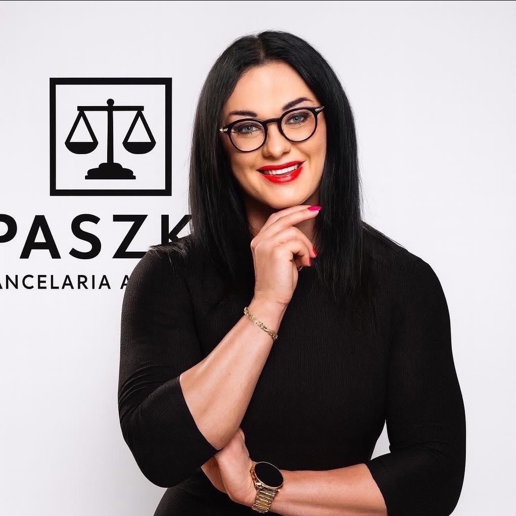 Portret uśmiechniętej kobiety w okularach na tle logo kancelarii prawnej z wagą. Kobieta w czarnej sukience, z zegarkiem i bransoletką. Profesjonalny wizerunek adwokata.