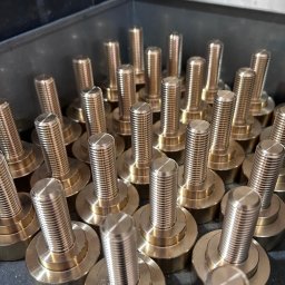 Inspirtech - Partia mosiężnych śrub z gwintem, ułożonych w szarym pojemniku. Widoczne detale obróbki i precyzyjne wykonanie gwintów. Metaliczny połysk i powtarzalność elementów.