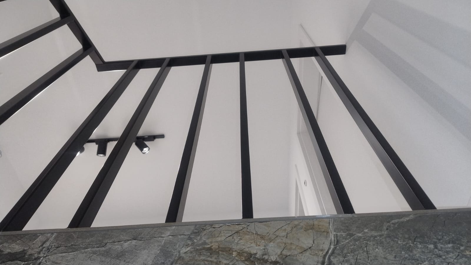 Nowoczesna balustrada schodowa z czarnych, metalowych prętów, widziana z dołu, z marmurowym wykończeniem podstawy i oświetleniem sufitowym w tle.