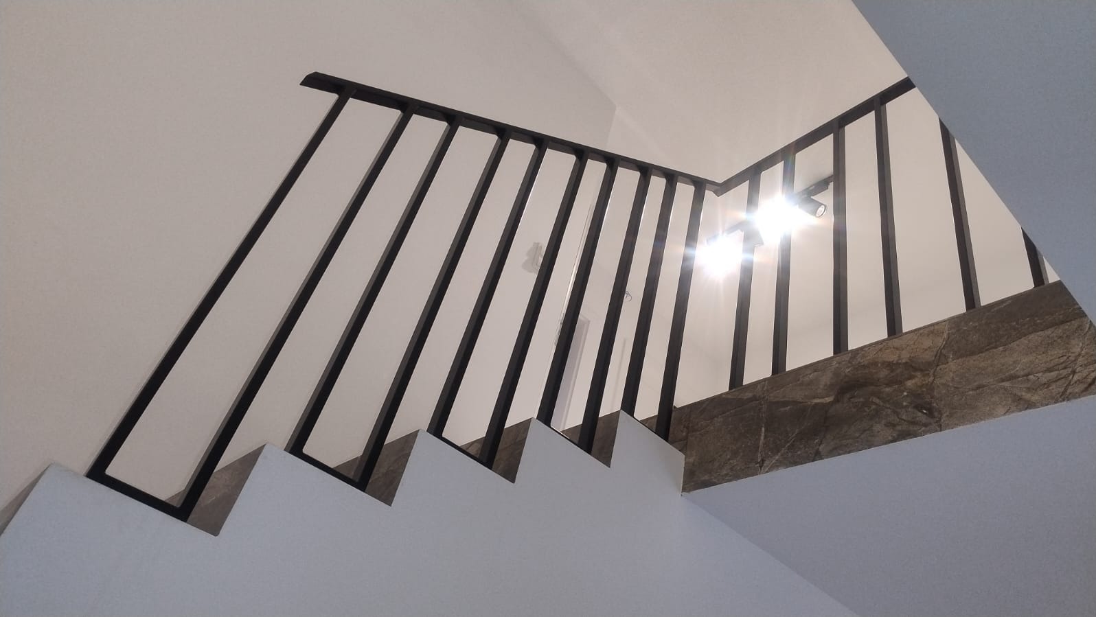Minimalistyczna, czarna balustrada schodowa z pionowymi szczeblami, zamontowana na białych stopniach. Widok z dołu, oświetlenie punktowe na suficie.