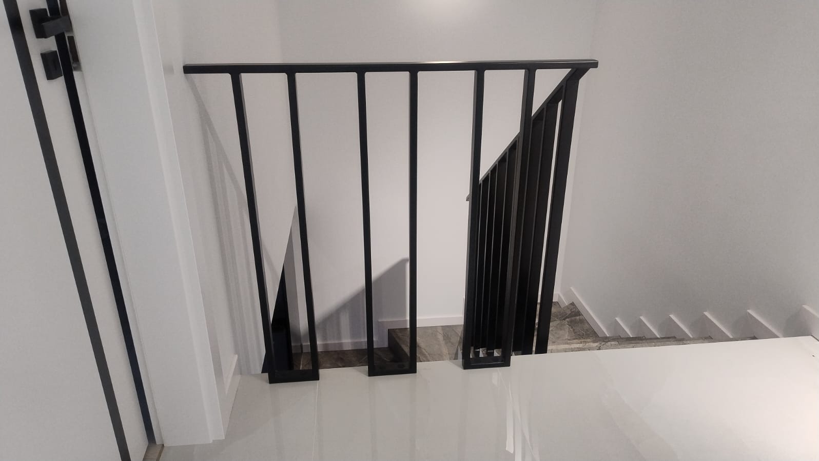 Minimalistyczna, czarna balustrada metalowa na schodach w nowoczesnym wnętrzu. Widoczne białe ściany i błyszcząca, jasna podłoga. Ujęcie z dołu, podkreślające geometrię konstrukcji.