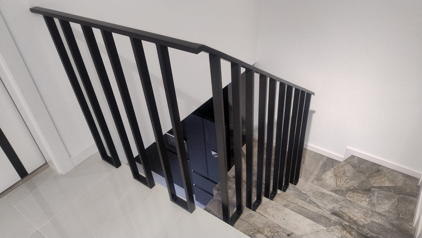 Nowoczesna, czarna balustrada schodowa z metalu, widziana z góry. Minimalistyczny design, jasne ściany, marmurowe schody. Geometryczne kształty poręczy.