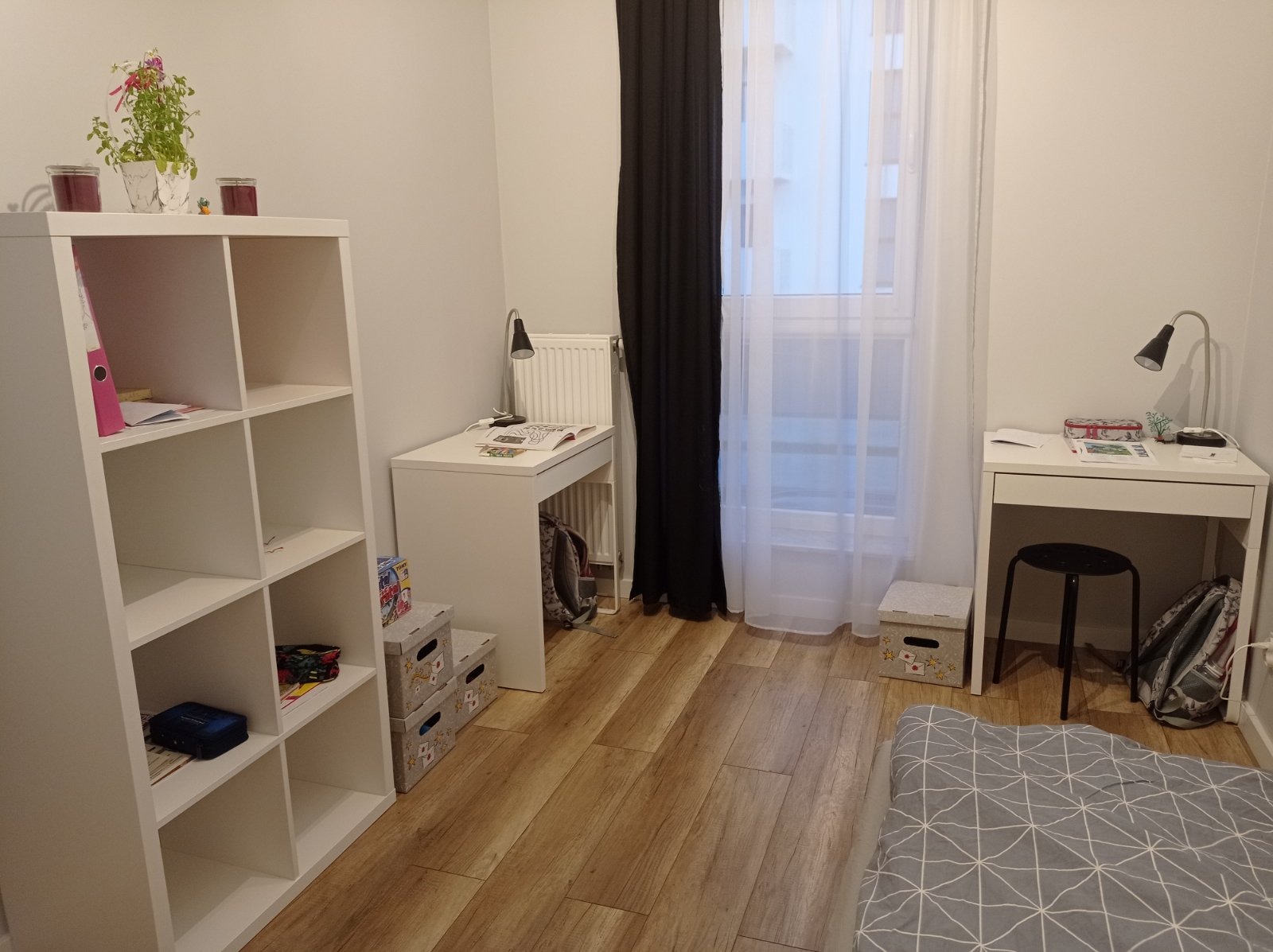 Pokój młodzieżowy po remoncie: panele podłogowe, białe meble (regał, biurka), okno z czarno-białymi zasłonami. Widok na fragment łóżka z geometrycznym wzorem.