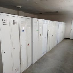 SkyDan - Rząd białych, metalowych szafek z numerami i naklejkami w szatni. Widoczne ślady użytkowania na szafkach i suficie. Perspektywa z dołu, oświetlenie sufitowe.