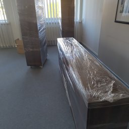 SkyDan - Meble zabezpieczone folią stretch w jasnym pomieszczeniu z oknami. Przygotowanie do transportu lub remontu. Ochrona przed uszkodzeniami i zabrudzeniami.