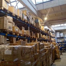 SkyDan - Magazyn z regałami wypełnionymi kartonami, oświetlony światłem dziennym z okien dachowych. Pracownik obsługuje wózek widłowy w wąskim korytarzu.